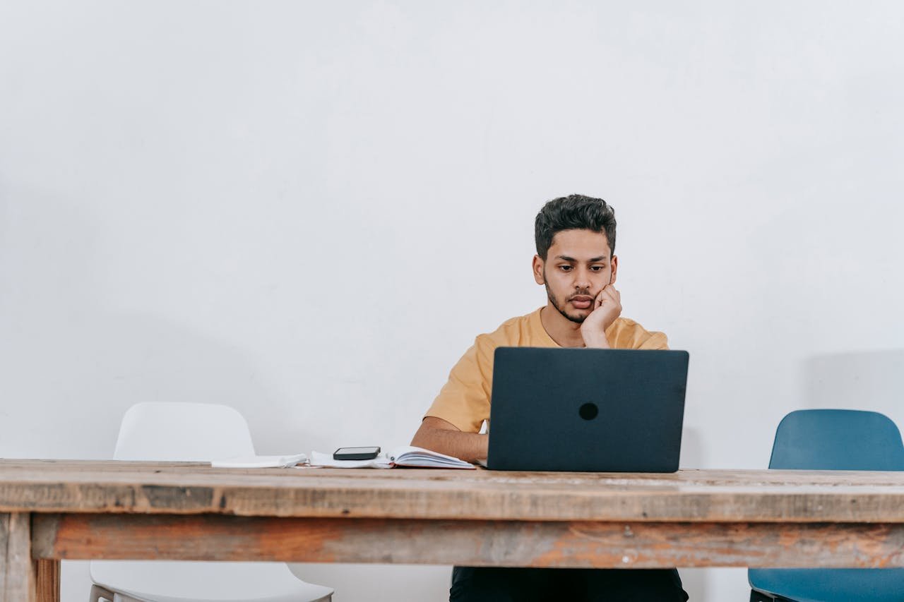 Focused young man studying on a laptop in a minimalist indoor workspace.