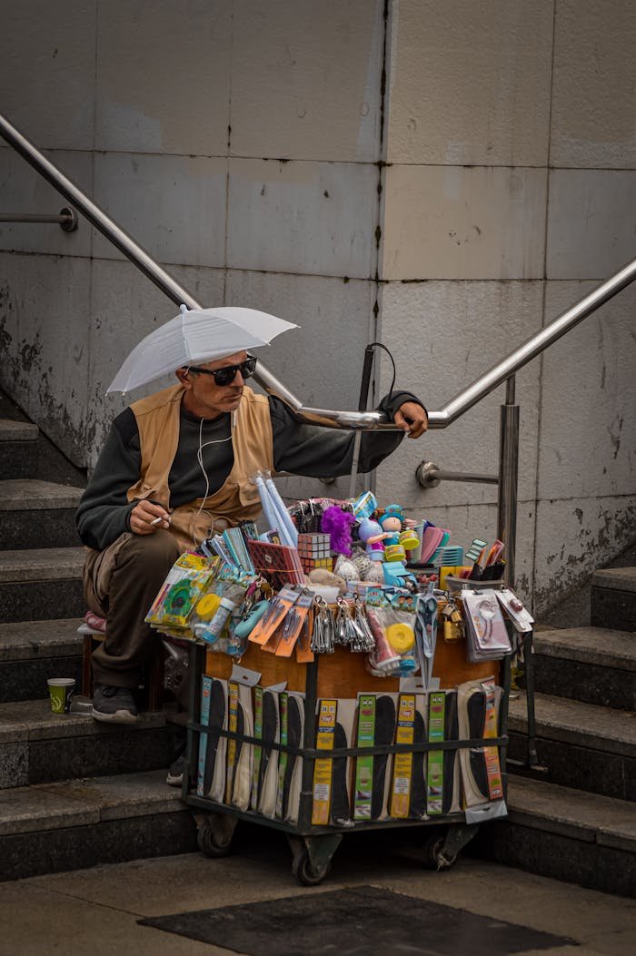 Urban street vendor with various items displayed on a cart, sitting by city steps.